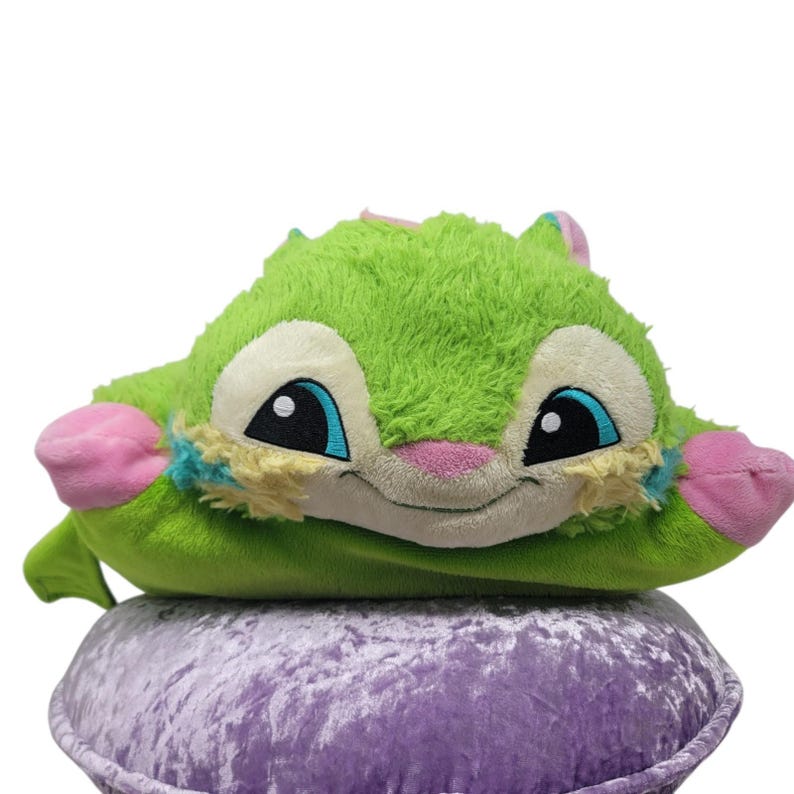 Puede incluir: Una almohada de felpa verde con forma de criatura con una cara sonriente, ojos azules y detalles en rosa. Descansa sobre una superficie de terciopelo morado. El dise&ntilde;o es de un personaje amigable.