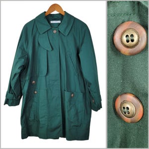 May include: A dark green trench coat with a classic design. The coat features a collared neckline, button closures, and two front pockets. The buttons are a marbled brown color. The coat is hanging on a wooden hanger.
