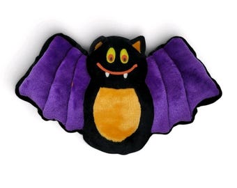 Puede incluir: Un juguete de peluche con forma de murciélago que presenta un cuerpo negro con una barriga naranja brillante, grandes alas moradas y una cara sonriente con ojos amarillos y dos pequeños colmillos blancos. El diseño destaca las alas y la cara.