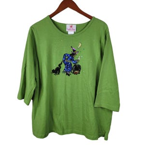 May include: Green long-sleeved t-shirt with a sequined image of a witch flying on a broomstick with a black cat and a cauldron. The text "Quacker Factory" is on the tag.