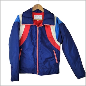 May include: A navy blue puffer jacket featuring a high collar, a bright red front zipper, and color-blocked panels in red, white, and light blue. The interior label reads "KOSSE by Guzzini". The jacket has long sleeves and two zippered side pockets.