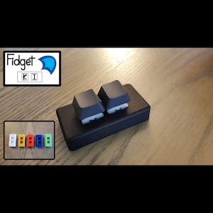 May include: Two black keyboard keys mounted on a black rectangular base. The base has a textured surface. The keys are labelled "K" and "I".
