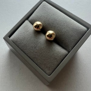 May include: A pair of gold-coloured, spherical stud earrings presented in a grey jewellery box with a soft, velvety interior. The earrings are highly polished, reflecting light, and are in a square, dark grey box.