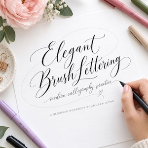 May include: A white practice workbook titled Elegant Brush Lettering modern calligraphy practice A BEGINNER WORKBOOK BY AMUSAN ILONA with black ink text, surrounded by several colored markers, a small dish with gold clips, and a pink flower.