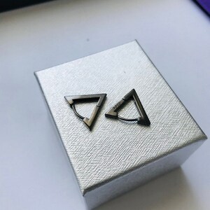 May include: A pair of dark grey triangular hoop earrings. The earrings are presented on a textured silver box. The earrings have a simple, geometric design. The earrings are made of metal with a matte finish. A modern accessory.