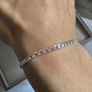 May include: A silver curb chain bracelet. The bracelet, made of a shiny metal, is worn on a wrist. The links are interlocked, forming a continuous pattern. The bracelet is a piece of jewellery, suitable for everyday wear.