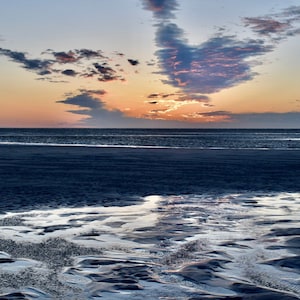 May include: A scenic view of a beach at sunset with a blue sky and pink clouds. The tide is out, revealing a network of shallow pools and wet sand.