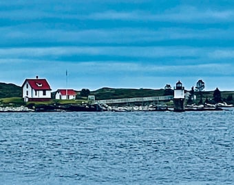 May include: A white lighthouse with a red roof sits on a small island with a white house and a wooden walkway. The lighthouse is surrounded by water and a blue sky with white clouds.