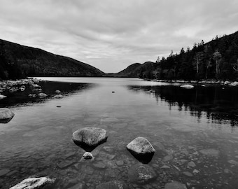 May include: A black and white photograph of a still lake with mountains in the background. The lake is calm and reflects the mountains. There are rocks in the foreground.