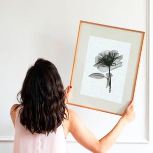 May include: A framed art print featuring a black and white floral illustration of a rose. The artwork is displayed in a gold-toned frame with a white mat. The image is held up against a white wall.