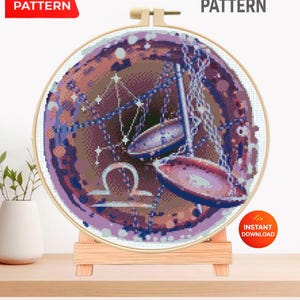 May include: A cross-stitch pattern featuring the Libra zodiac symbol, displayed in a wooden embroidery hoop. The design uses shades of purple, brown, and blue. Text on the image reads "PDF PATTERN" and "CROSS STITCH PATTERN".