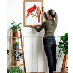 May include: A framed watercolor print of two red cardinal birds perched on a branch. The artwork features a male and female cardinal with detailed feather textures and a naturalistic style. The frame is a light brown color.