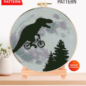 Puede incluir: Un patrón de punto de cruz en un aro de madera con una silueta negra de un T-Rex en bicicleta frente a un cielo iluminado por la luna. El texto "CROSS STITCH PATTERN" y "PDF PATTERN" son visibles. El diseño se muestra en un soporte de madera.