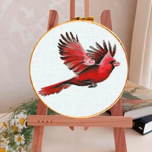 May include: A cross-stitch embroidery of a red cardinal bird in flight, set within a round wooden hoop. The bird's wings are spread, showcasing detailed stitching. The embroidery is displayed on a small wooden easel.