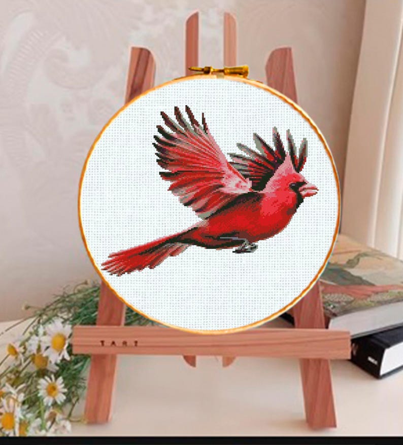 May include: A cross-stitch embroidery of a red cardinal bird in flight, set within a round wooden hoop. The bird's wings are spread, showcasing detailed stitching. The embroidery is displayed on a small wooden easel.