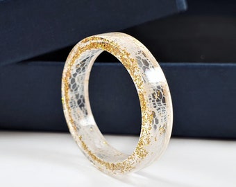 May include: A circular ring made of clear resin featuring an embedded white lace pattern and fine gold glitter particles throughout the band. The ring is positioned upright in front of an open dark blue jewelry box with a black interior lining.
