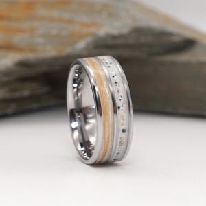 Op de afbeelding: Een zilverkleurige metalen ring met een centrale inleg bestaande uit een lichtbruine houten strook naast een gespikkelde witte materiaalband. De ring heeft een gepolijste metalen afwerking en een glad, afgerond buitenprofiel.