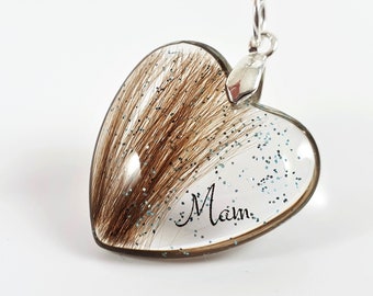 May include: A silver heart-shaped pendant necklace with a clear resin insert containing brown hair and blue glitter. The word "Mam" is inscribed on the pendant.