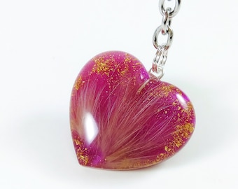 May include: A pink heart-shaped keyring with gold glitter embedded in clear resin. The heart contains a delicate, white feather inside.