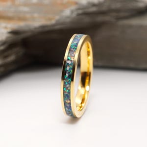 May include: A gold-colored ring with a band of iridescent blue, green, and white stones. The ring is set against a white background with a gray stone in the background. The ring is a piece of jewelry.