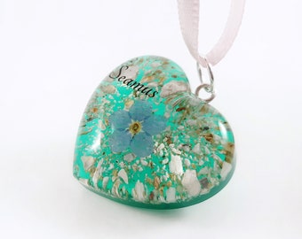 May include: A teal heart-shaped pendant with a forget-me-not flower and the name "Seamus" inscribed on it. The pendant is on a pink ribbon.