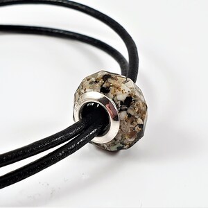 May include: A close-up of a silver charm bracelet with a faceted bead filled with sand and shells. The bead is attached to a black leather cord.