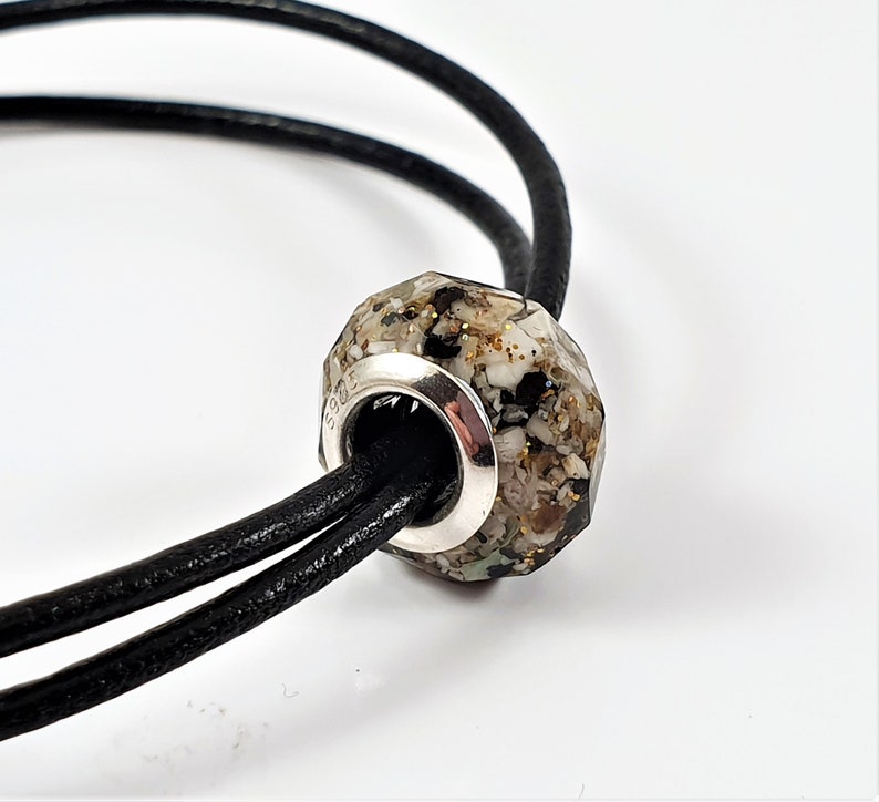 May include: A close-up of a silver charm bracelet with a faceted bead filled with sand and shells. The bead is attached to a black leather cord.