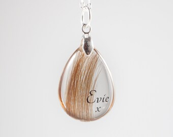 May include: A silver teardrop-shaped pendant necklace with a clear resin insert containing brown hair and the inscription "Evie x".