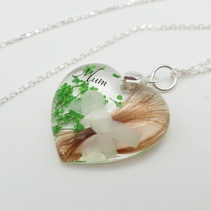 May include: A heart-shaped pendant necklace with a silver chain. The clear resin pendant contains brown hair, green sprigs, and a white flower. The word "Mum" is written in black script on the pendant.