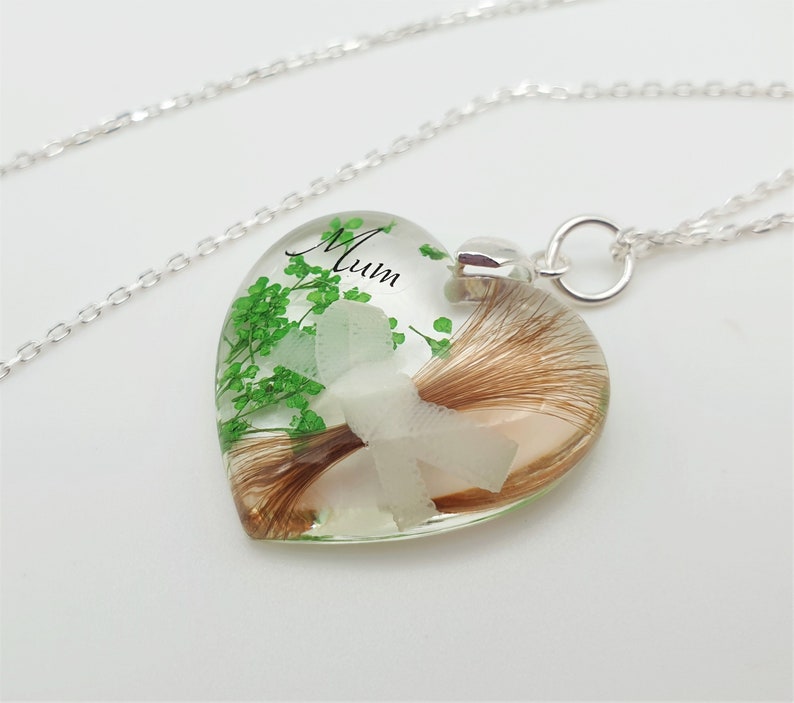 May include: A heart-shaped pendant necklace with a silver chain. The clear resin pendant contains brown hair, green sprigs, and a white flower. The word "Mum" is written in black script on the pendant.