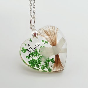 May include: A heart-shaped pendant with a silver chain. The clear resin heart contains brown hair, a white ribbon, green clovers, and the word "Mum" in black script. The pendant is suspended from a silver bail.