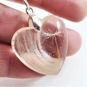 May include: Heart-shaped pendant with a clear resin body. The pendant contains white fibers, a dandelion seed, and pink glitter. The pendant has a silver-colored bail.