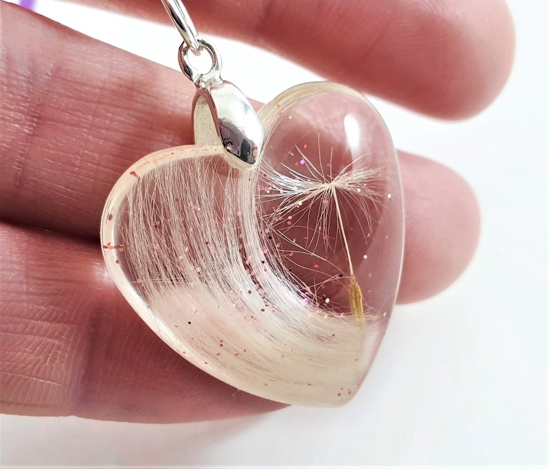 May include: Heart-shaped pendant with a clear resin body. The pendant contains white fibers, a dandelion seed, and pink glitter. The pendant has a silver-colored bail.