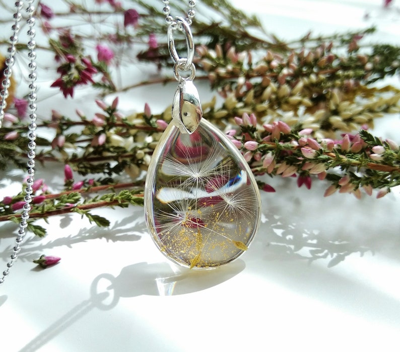 May include: A silver chain necklace with a teardrop-shaped pendant. The pendant is filled with a clear resin and contains a dried dandelion with gold flecks.