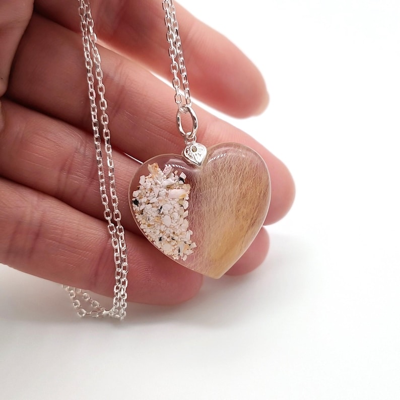 May include: A silver chain necklace with a heart-shaped pendant. The pendant is filled with clear resin and contains a mixture of sand and fur.