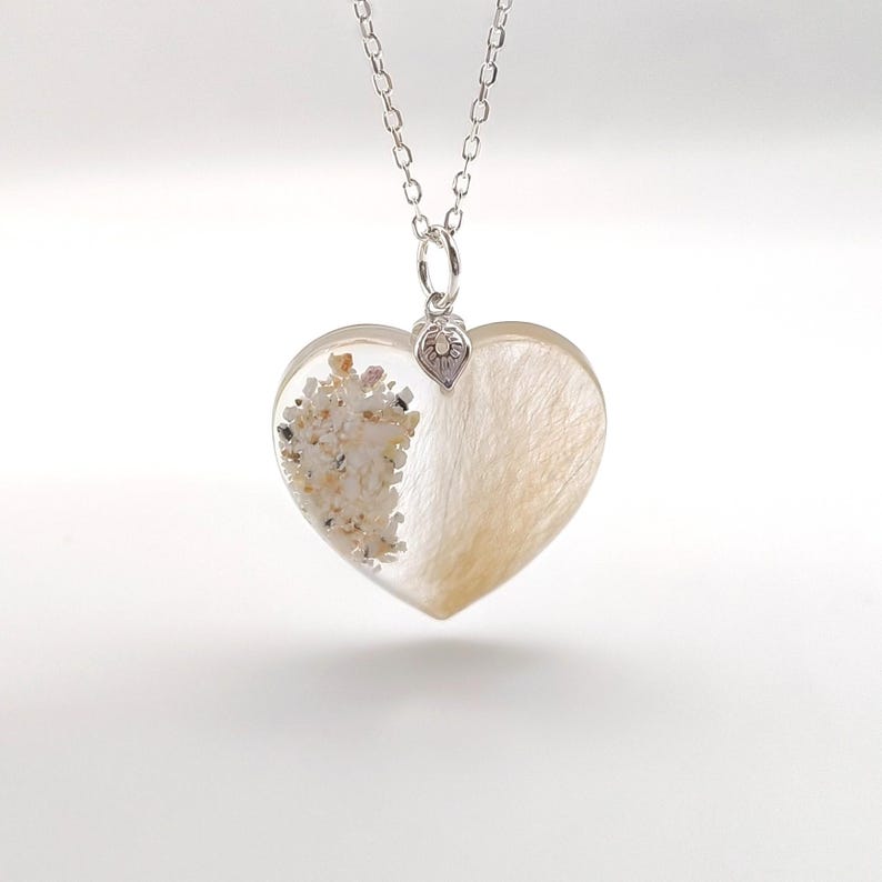 May include: A silver chain necklace with a heart-shaped pendant. The pendant is filled with clear resin and contains sand and hair. A small silver heart charm hangs from the pendant.