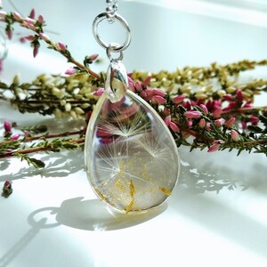 May include: A silver chain necklace with a teardrop-shaped pendant. The pendant is filled with clear resin and contains a dried dandelion with gold glitter.