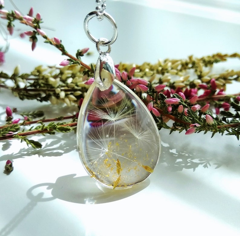 May include: A silver chain necklace with a teardrop-shaped pendant. The pendant is filled with clear resin and contains a dried dandelion with gold glitter.