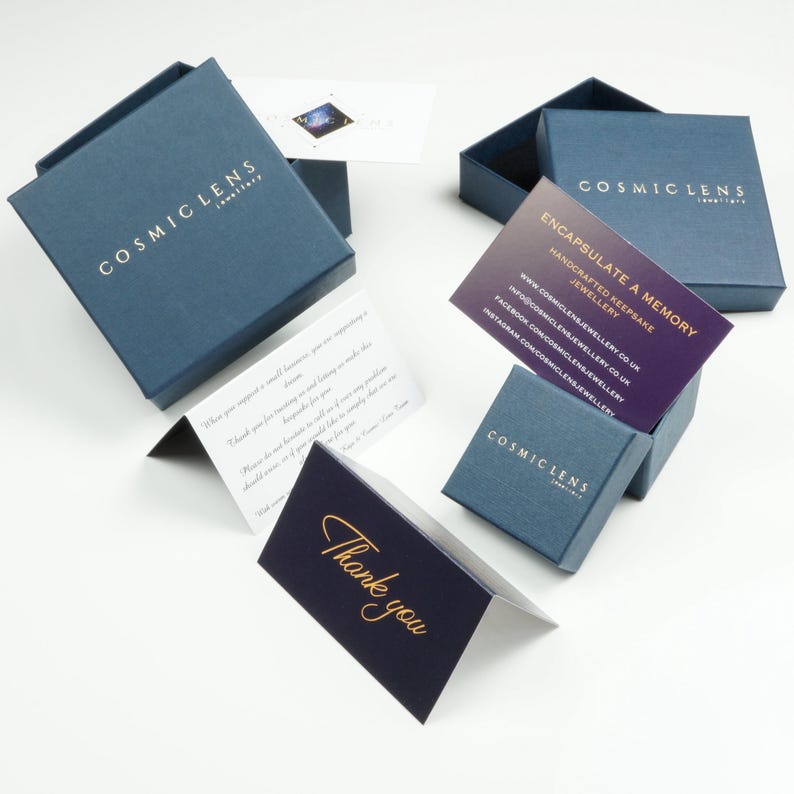May include: Several open navy blue jewelry boxes with gold lettering reading "COSMIC LENS Jewellery".  Cards with text, including "Encapsulate a Memory" and "Thank you" are displayed. The boxes and cards are on a white surface.