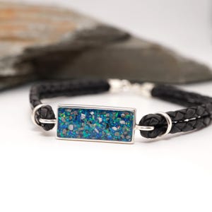 May include: A black leather bracelet with a rectangular, silver-toned centerpiece. The centerpiece is filled with a mosaic of blue, green, and white stones. The bracelet has a braided design and a silver clasp.