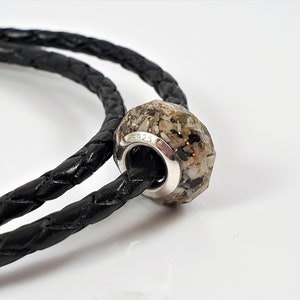 May include: A silver charm bracelet with a black leather cord and a faceted glass bead filled with sand and small shells. The bead is marked with the number "S925".