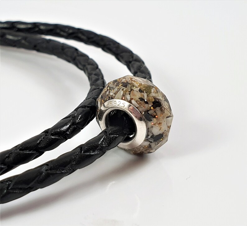 May include: A silver charm bracelet with a black leather cord and a faceted glass bead filled with sand and small shells. The bead is marked with the number "S925".