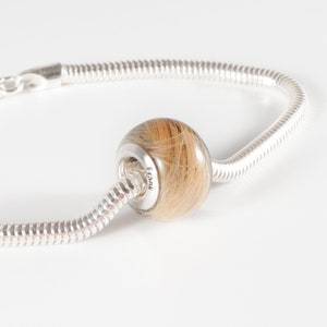 May include: A silver charm bracelet with a round bead filled with brown hair. The bead is engraved with the name "Frank".