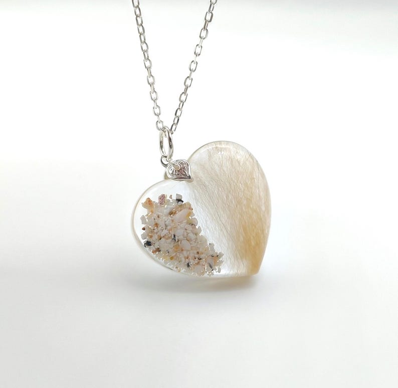 May include: A silver chain necklace with a clear heart-shaped pendant. The pendant is filled with sand and hair, creating a unique and sentimental piece of jewelry.