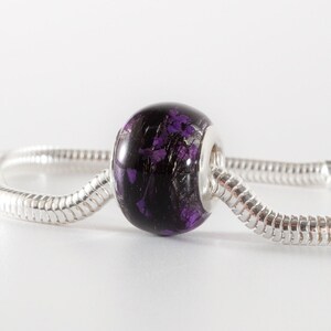 May include: A round, dark purple bead with embedded purple floral elements and fine black fibers, featuring a silver-toned metal core. The bead is threaded onto a flexible, segmented silver-toned snake chain bracelet with a polished finish.
