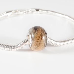 May include: A silver charm bracelet with a round bead filled with human hair. The bead is a light brown color with dark brown streaks.