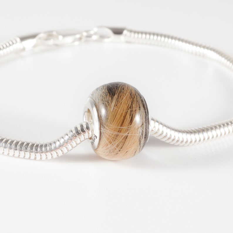 May include: A silver charm bracelet with a round bead filled with human hair. The bead is a light brown color with dark brown streaks.