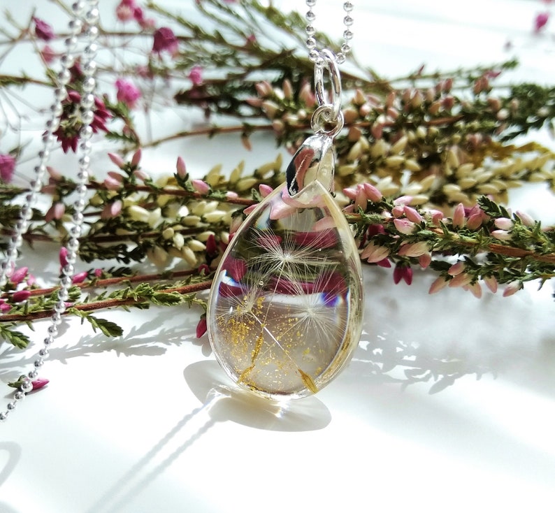 May include: A silver chain necklace with a teardrop-shaped pendant. The pendant is filled with clear resin and contains a dried dandelion with gold glitter.