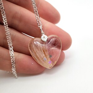 May include: A heart-shaped pendant necklace with a silver chain. The clear resin pendant contains strands of brown hair, a dandelion seed, and small star-shaped glitter. The pendant is held by a silver bail.