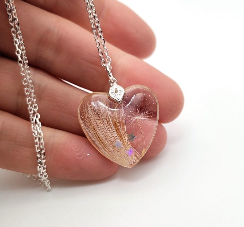 May include: A heart-shaped pendant necklace with a silver chain. The clear resin pendant contains strands of brown hair, a dandelion seed, and small star-shaped glitter. The pendant is held by a silver bail.